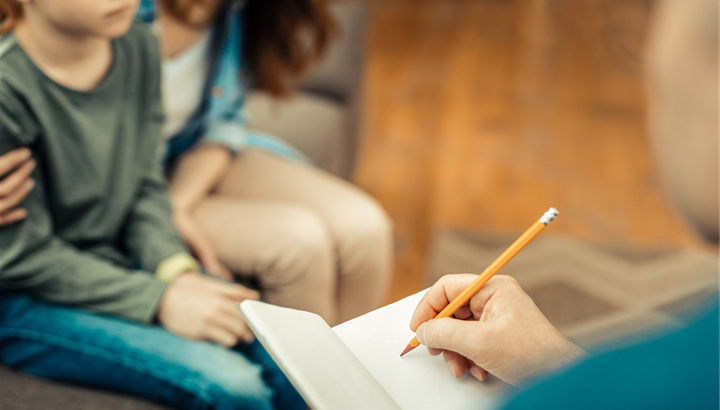 close up of person writing in a notebook, mom and son in background, mom embracing the child