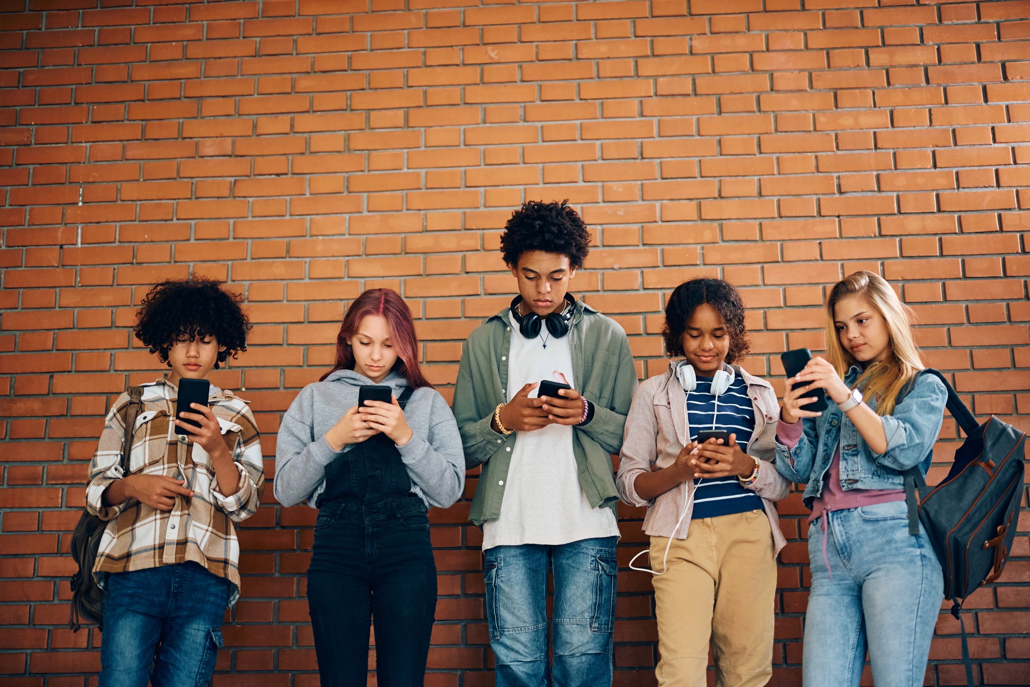 Five teens standing against a brick wall looking at their phones