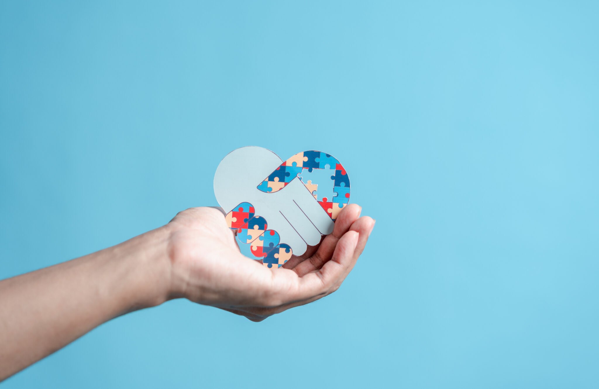 close up of hand with cutout of 2 hands in a handshake in the shape of a heart and one is made of colorful puzzle pieces