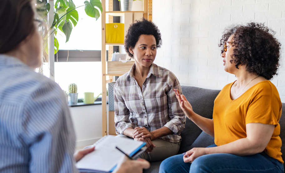 two women sitting on couch, one talking, the other listening in therapist office and therapist taking notes