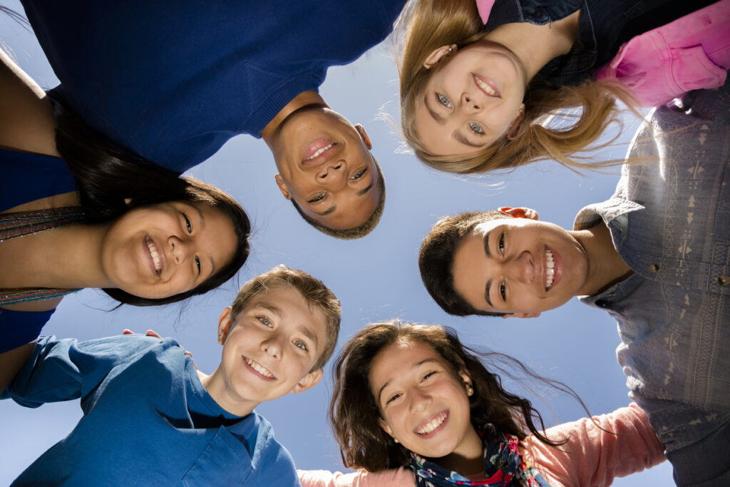 Multi-ethnic group of teenage friends hang out, huddle together with arms around each other outdoors. Blue sky background.
